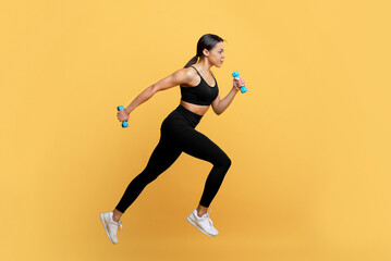 Wall Mural - Active lifestyle. Fit black lady running with two dumbbells in hands, working out on yellow studio background