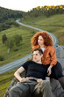 Relaxed Caucasian Couple Sitting On The Head Of Mountain, Having Rest After Climbing, Looking At Side In Contemplation of Nature Landscape Around. Road In The Background. Man And Redhead Woman