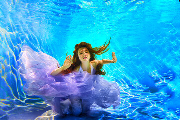 Girl in a beautiful dress under blue water. Female model posing under water in swimming pool