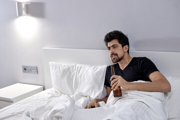 Indoor shot of grumpy young male relaxing in bedroom, watching television series, drinking alcohol in bed, commenting what happening on screen. bearded bachelor male at home have rest