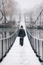 A Woman Walks Over The River On A Suspension Bridge In Winter Day. Young Girl In Warm Clothes Stands On A Wooden Footbridge In Cold Snowy Day