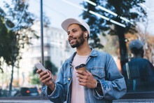 Smiling Spanish Hipster Guy In Bluetooth Headphones Enjoying Positive Music Playlist From Mobile Radio Using 4g Wireless In City, Millennial Male Tourist In Earbuds Holding Coffee To Go And Smartphone
