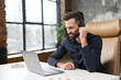 © Vadim Pastuh - Concentrated young guy in smart casual wear is using headset and laptop for online communication, supporting, selling. Guy sitting at the office desk