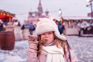 Russia Moscow People walk on the red square, a little funny girl eats a gingerbread on a stick against the background of a Christmas fair and a carousel in winter. High quality FullHD footage