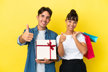 Wall Mural - Young couple holding shopping bags and present isolated on yellow background giving a thumbs up gesture with both hands and smiling