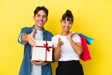 Wall Mural - Young couple holding shopping bags and present isolated on yellow background shaking hands for closing a good deal