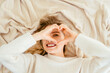 © Iryna - Above view smiling rested lovely blond female looking at camera through shape heart fingers feels refreshed starting new day, good morning, time to get up concept.