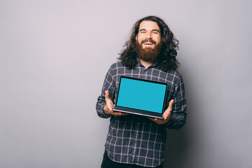 Cheerful young bearded hipster man showing blank laptop screen over grey background.