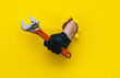 © shchus - A righ hand in a black knitted glove holds a red monkey wrench. Torn hole in yellow paper. The concept of a worker, labor migrant, a master of his craft. Copy space.
