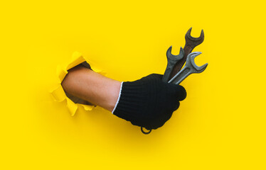 A hand in a black cloth knitted glove holds three wrench. Torn hole in yellow paper. The concept of a worker, labor migrant, a master of his craft. Copy space.