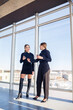 © DSMT - Two successful female managers talking about a joint project while standing by the window in the office. Young women economists, dressed in formal attire, talk during their breaks and drink coffee.