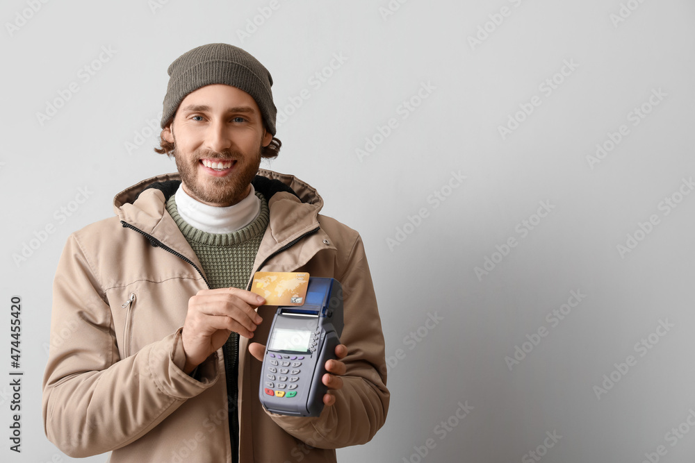 Young man with banking terminal and credit card on light background