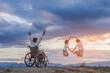 © AungMyo - Woman Sitting in wheelchair raised hands and children jumping on mountain at sunset background. Group of Happy Family concept.