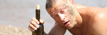 Drunk Young Man In Sand Lying On Beach With Bottle Of Wine In His Hands
