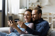 Happy millennial married couple using credit card, smartphone for shopping from home, making payment for purchase from virtual wallet on online banking service, financial application