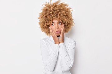 Wall Mural - Portrait of sad bored woman keeps hands on cheeks attends boring meeting rolls eyes has curly blonde hair dressed casually isolated over white background grimaces displeased feels indifferent