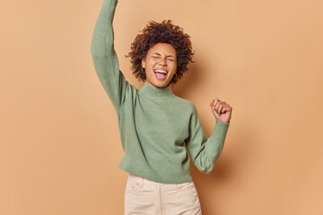 Wall Mural - Horizontal shot of overjoyed curly haired woman dances with excitement shakes arms moves actively laughs out dressed in casual jumper and trousers isolated over beige background. Joy concept