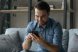 © fizkes - Smiling young man sit on couch with smartphone, choose goods buy services online, chatting in social media, single guy enjoy e-dating e-services, new mobile application spend free time alone at home