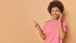 © WHstudio Leushin N - Studio shot of happy pleased young woman has telephone conversation discusses something indicates index finger at blank copy space makes phone call isolated over beige background copy space area