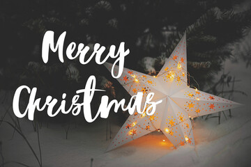 Wall Mural - Merry Christmas greeting card. Merry Christmas text on big Christmas star glowing at snowy pine tree. Seasons greeting. Happy Holidays! Handwritten sign