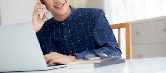 Wall Mural - Young asian man talking phone and work from home with laptop computer, freelance using notebook to internet online and speak on smartphone, domestic life, business and communication concept.