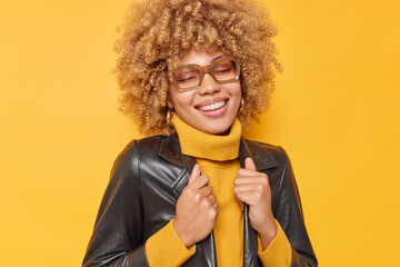 Wall Mural - Photo of positive young European woman with curly hair dressed in stylish clothes recalls nice memories keeps eyes closed isolated over yellow background. People clothes and emotions concept