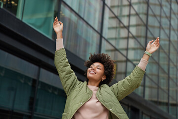 Wall Mural - carefree joyful woman dressed in casual jacket raises arms poses against modern glass buildings in d