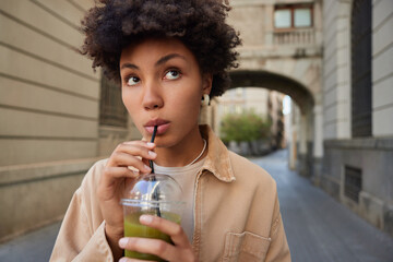 Wall Mural - Organic food and weightloss drinks concept. Thoughtful curly haired woman drinks fresh raw detox vegetable smoothie has vegetarian meal looks pensively forward strolls in city leads healthy lifestyle
