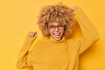 Wall Mural - Positive woman with curly bushy hair has fun and dances carefree keeps raised up moves actively laughs out wears spectacles casual turtleneck isolated over yellow background. Happy emotions.