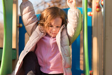 Small Child With Long Red Hair Against The Background Of A Blue Sky In Warm Clothes On The Playground