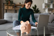 Close up of piggy bank on table, happy Hispanic woman manage household budget pay online on laptop from home. Provident economical Latin female make payment on internet on computer. Saving concept.
