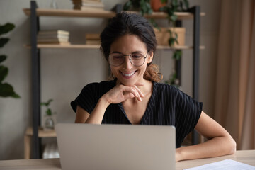 Sticker - Smiling young Latino woman in glasses look at laptop screen work online on gadget at home office. Happy Hispanic female in eyewear study on computer distant, talk on webcam video call on device.