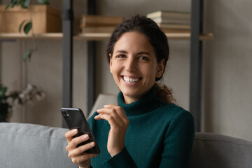 Poster - Portrait of happy millennial Latino woman relax on couch at home use modern cellphone gadget texting messaging online. Smiling young Hispanic female client or user browse web on smartphone device.