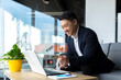 © Liubomir - Asian boss transfers money from a bank card, sitting in the office, satisfied with the result businessman smiles, works on laptop