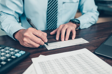 Business man hand writing and signing white blank bank check book and dollar bill, coin, laptop and graph chart on the desk at office. Payment by check, paycheck, payroll concept.