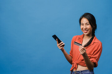 Wall Mural - Young Asia lady using phone and credit bank card with positive expression, smiles broadly, dressed in casual clothing and stand isolated on blue background. Happy adorable glad woman rejoices success.