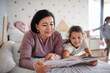 © Halfpoint - Happy small girl with senior grandmother indoors in bedroom at home, reading on bed.