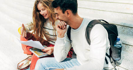 Wall Mural - Horizontal image of a two young university students studying sitting on the stairs of the college campus. Woman and man sitting outside learning together from the books.