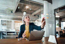 Senior Businesswoman Putting Adhesive Note On Glass Wall