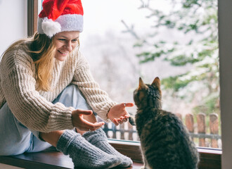 Wall Mural - Young cheerful girl in a Sanat Claus hat sitting at home on the windowsill in a warm sweater playing with a gray cat, Christmas and new year concept