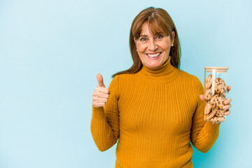 Middle age caucasian woman holding cookies jar isolated on blue background smiling and raising thumb up