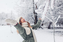 Happy Young Woman Playing With Snow In Snowy Winter Park Wearing Warm Knitted Clothes And Having Fun.