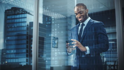 Poster - Portrait of Successful Black Businessman Wearing Suit Standing, Using Smartphone Looking out of the Window. Successful African CEO Planning e-Commerce Investment Strategy. From Outside Shot