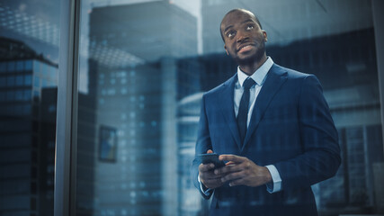 Poster - Portrait of Successful Black Businessman Wearing Suit Standing, Using Smartphone Looking out of the Window. Smiling Stock Market Investment Manager Working Closing the Deal. Shot from Outside