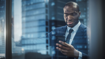 Poster - Portrait of Successful Black Businessman Wearing Suit Standing, Using Smartphone Looking out of the Window. Smiling Stock Market Investment Manager Working Closing the Deal. Shot from Outside