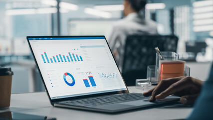 Successful Black Businessman Sitting at Desk Working on Laptop Computer in Big City Office. Hard Working Top Manager Doing Research for e-Commerce Project with Graps, Charts and Tables. Over Shoulder