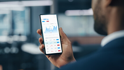Black Businessman Holding Smartphone and Checking Stock and Cryptocurrency Market in Office. African-American Businessperson using Internet with Mobile Phone Device. Over Shoulder Shot