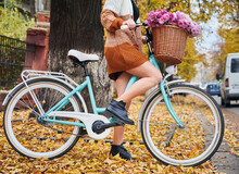 Female Bicyclist In Knitted Cardigan Riding Bike With Bouquet Of Flowers In Basket. Stylish Young Woman With Bicycle Standing On The Street With Yellow Autumn Leaves.