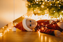 Sad Serious Baby In Santa Hat, Lying On The Floor, On A Christmas Tree With Lights Background, Looking At Camera.