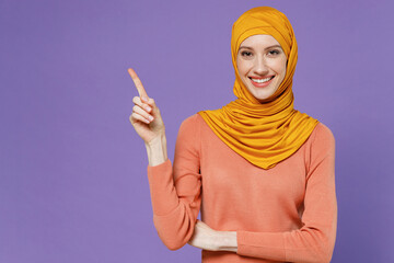 Wall Mural - Promoter smiling fun young arabian asian muslim woman in abaya hijab yellow clothes pointing away on workspace area copy space mock up isolated on plain pastel light violet background studio portrait.